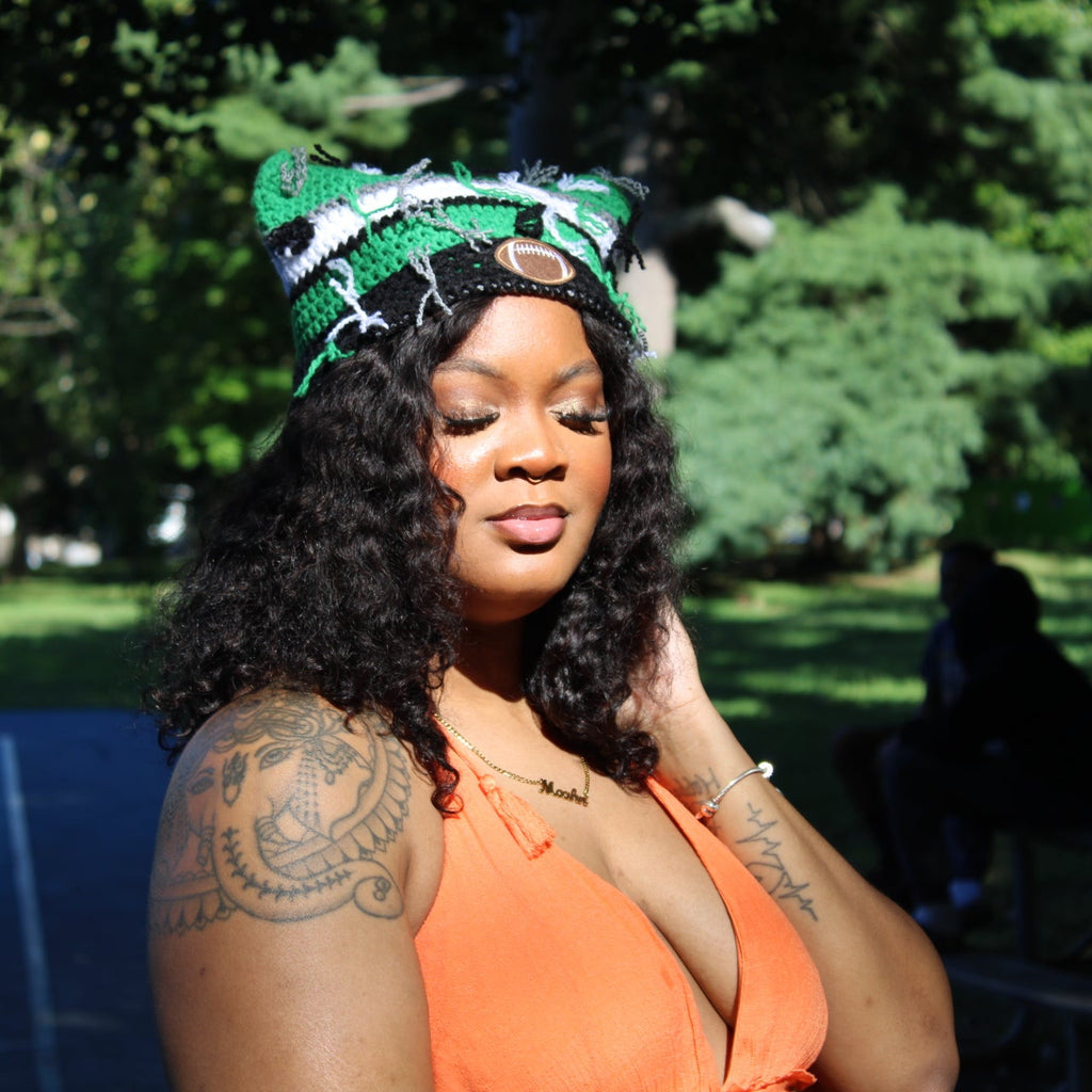 A woman outdoors in a park wearing a green and white patterned knit headband with a small gold circular accent, paired with a bright orange sleeveless top. She has curly black hair and visible tattoos on her arm.