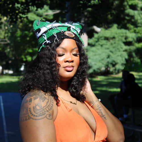 A woman outdoors in a park wearing a green and white patterned knit headband with a small gold circular accent, paired with a bright orange sleeveless top. She has curly black hair and visible tattoos on her arm.