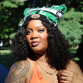 A woman outdoors in a park wearing a bright orange halter top and a green, white, and black patterned headband, with a tattoo visible on her upper arm.