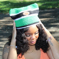 A woman outdoors wearing a crocheted hat with green, black, white, and brown horizontal stripes, holding the hat in place with both hands.