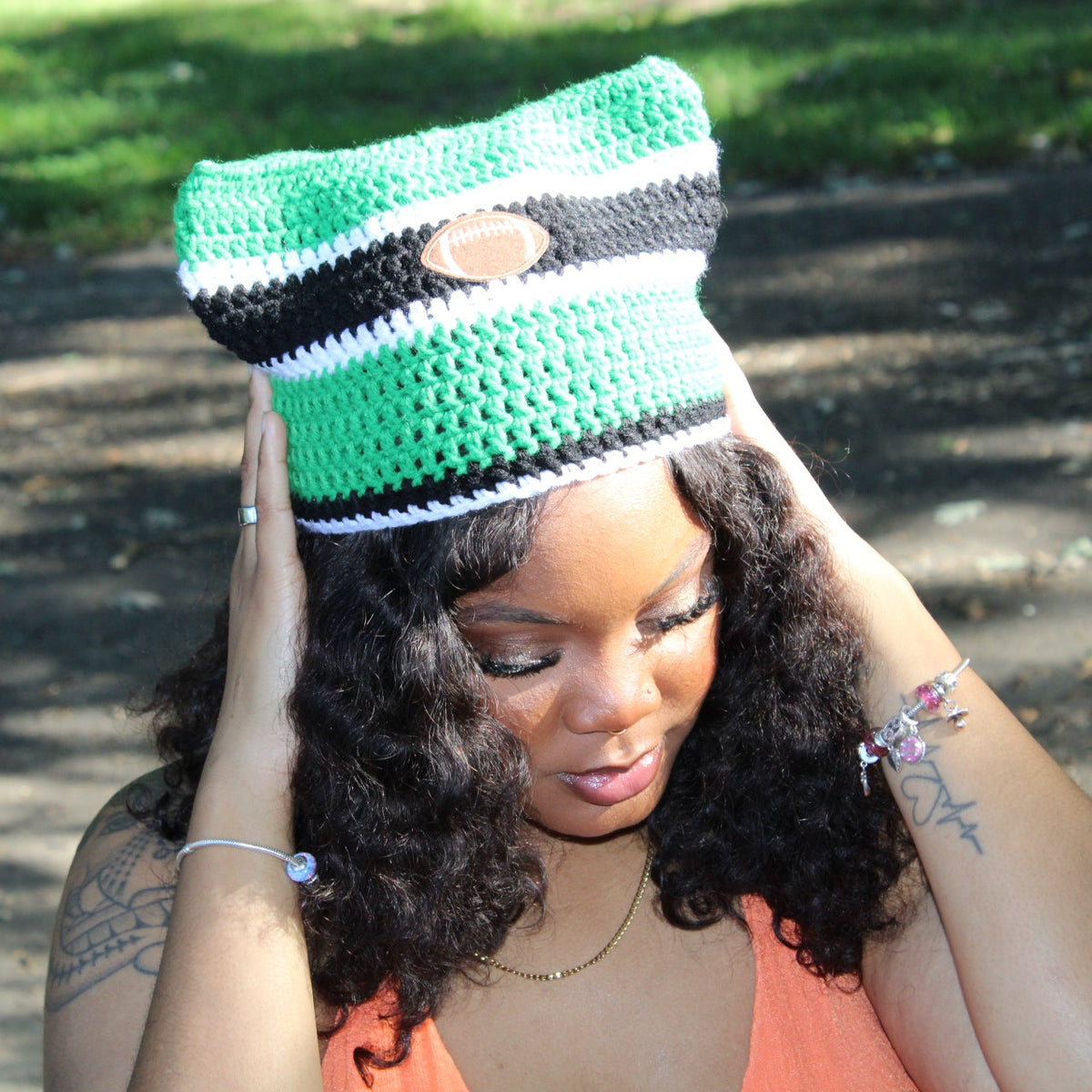 A woman outdoors wearing a crocheted hat with green, black, white, and brown horizontal stripes, holding the hat in place with both hands.