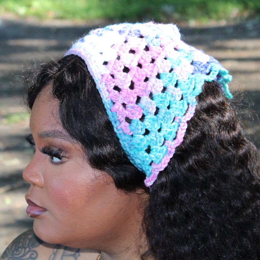 A woman in profile wearing a crocheted headband with a pastel pink, purple, and light blue color gradient, photographed outdoors on a path with grass in the background.