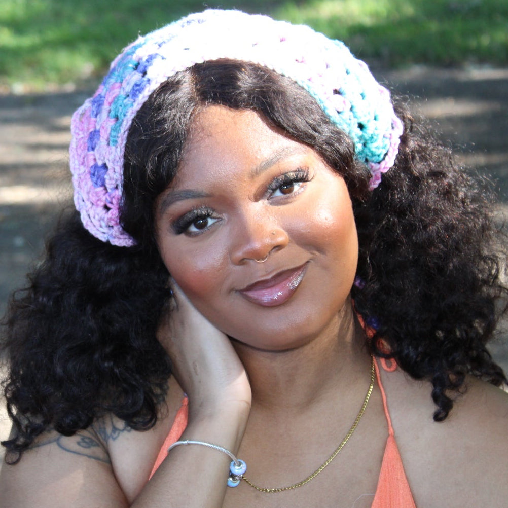 A woman outdoors wearing a crocheted headband in pastel pink, purple, and blue, paired with a peach-colored halter top. The background shows a blurred dirt path and green grass under natural daylight.