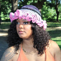A woman wearing a handmade crocheted hat with a pink ruffle trim and a striped pattern of purple, black, and white, photographed outdoors in a park.