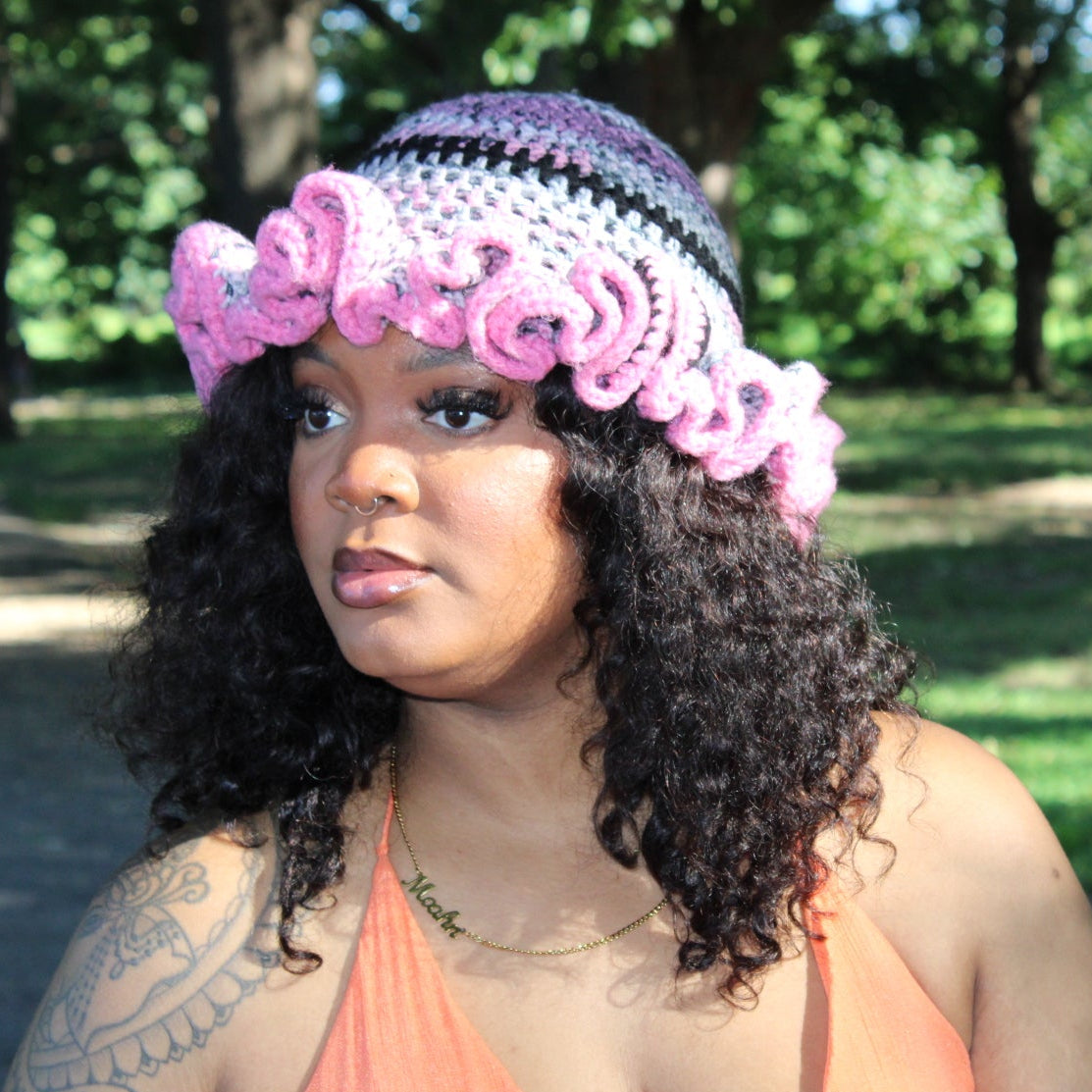 A woman wearing a handmade crocheted hat with a pink ruffle trim and a striped pattern of purple, black, and white, photographed outdoors in a park.