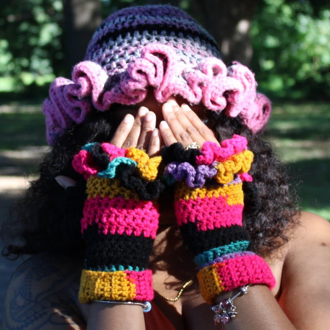 A person outdoors in a park wearing a colorful crocheted hat and matching fingerless gloves, with their hands covering their face. The background shows trees and a park path.