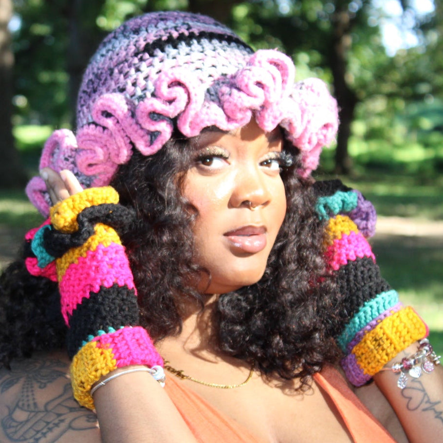 A woman outdoors wearing a handmade multicolor crochet hat with pink ruffles, matching striped crochet gloves, and a peach-colored top, posing in a park with trees in the background.