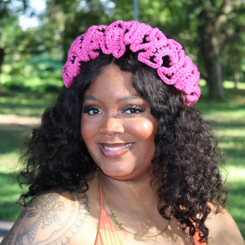 A smiling woman with curly black hair wearing a bright pink crocheted headband, standing outdoors in a park with trees and grass in the background.