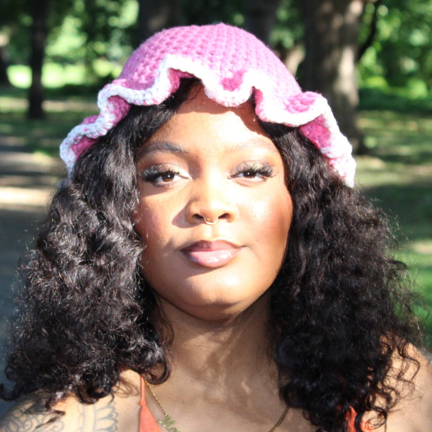 A woman with curly black hair wearing a pink crocheted hat with a ruffled brim, standing outdoors on a park path with trees in the background.