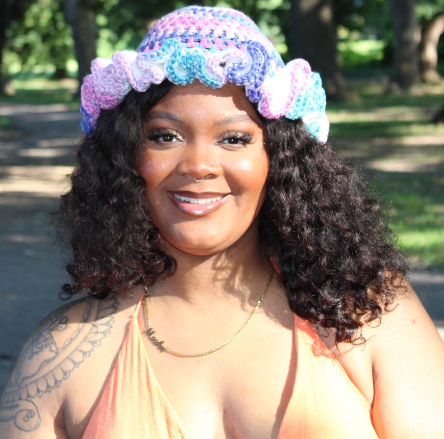 A smiling woman outdoors wearing a colorful crocheted hat with pink, purple, and blue flowers, standing next to a tree in a park.