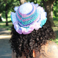 A person photographed from behind outdoors on a tree-lined path, wearing a crocheted sun hat with ruffled trim in pastel blue, purple, pink, and white.