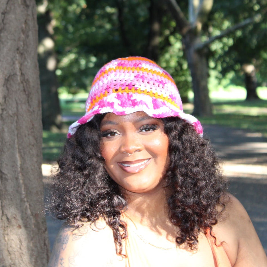 A smiling woman with curly dark hair stands outdoors next to a large tree trunk in a park, wearing a colorful knit bucket hat with pink, orange, and white patterns.