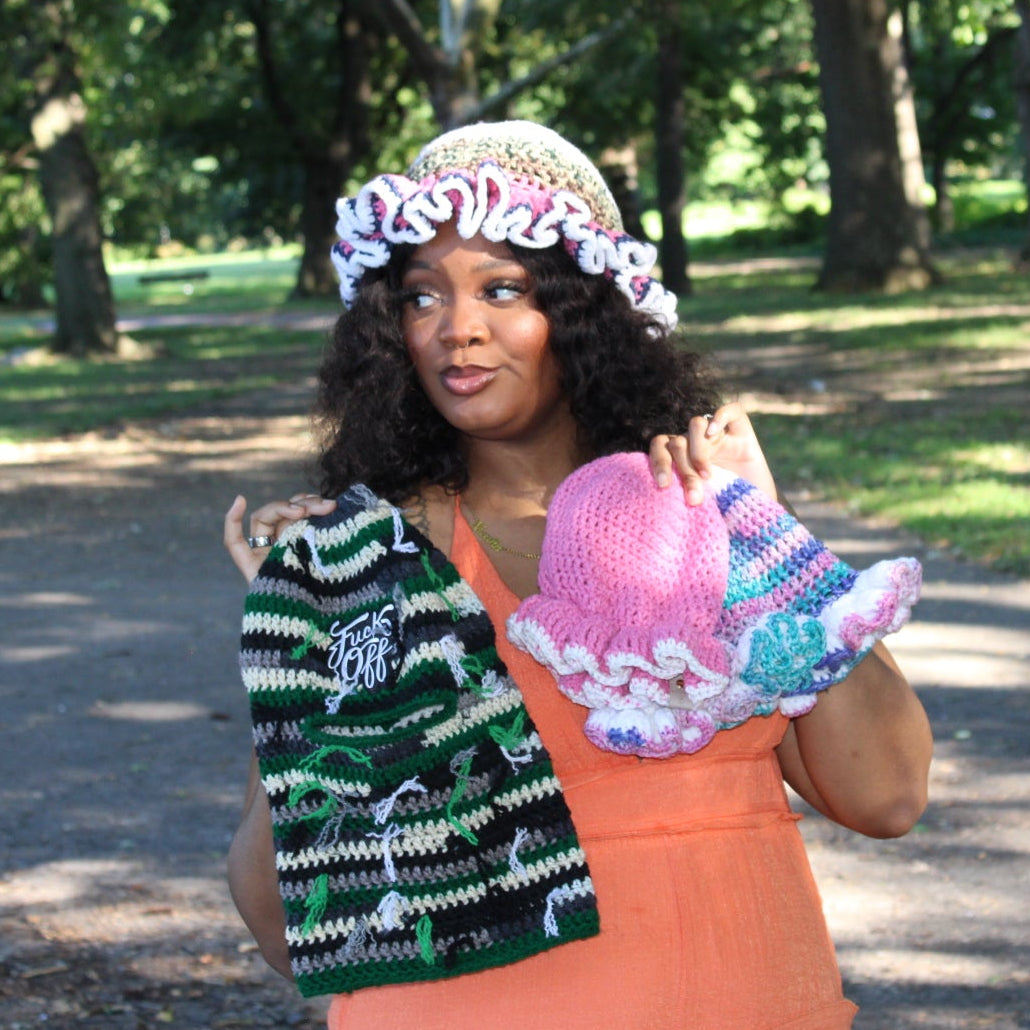 A woman standing in a park holding a crocheted green and black striped hat and a crocheted pink and blue hat, wearing a peach-colored dress and a beige and white ruffled hat.